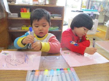 『お絵かきたのしいな』 　普段から、友だちと「それ、かわいいね」「わたしも、かいてみよう」などお話をしながらお絵かきを楽しんでいます。これは冬の絵をプラバンに油性ペンで描きました。「つるつるしてる」と紙との違いに気付いたり、「とうめいになっている」と透き通ることに気付く子どもたちでした。窓に貼ると「かげもにじいろや」「ステンドグラスみたい」と気付きがいっぱいでした。