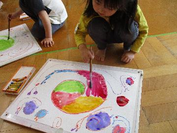 『おいしいケーキ、いかがですか？』 　濃度の濃い、ぽってりとした絵の具にバニラエッセンスを入れて、ケーキを描きました。「わあ、いいにおい」「なにをトッピングしようかな」と友だち同士でお話ししながらとても美味しそうなケーキが出来上がりました。描きたいものによって、保育士が絵の具の濃さを調節します。「きょうのえのぐはちょっとこいな」「きょうははサラサラやな」などの気付きも大切にしています。