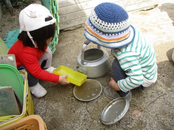 『おりょうりできるかな？』 やかんに水や葉っぱ、砂を入れて「おちゃどうぞ～」と話しながら遊んでいます。食べる前に「てをあらってきて」と言いながら友だちや保育士とごっこあそびをして楽しみます。